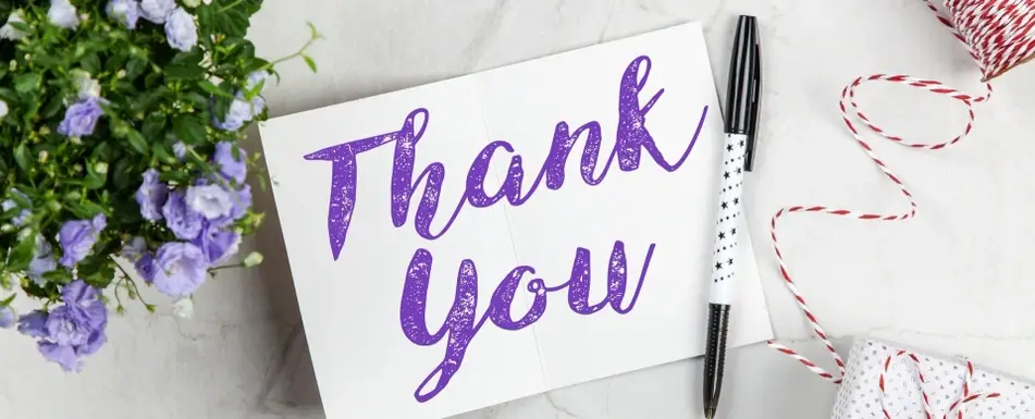 A thank you letter sitting on a table with the words "Thank you" written in purple on a white cardstock, with flowers and a roll of red and white twine nearby.