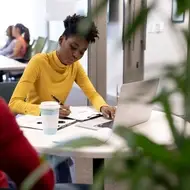 A Black student wearing a bright yellow turtle neck studies at a round table in the library.